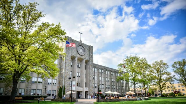 A college building with a clock tower, American flag, and trees in the foreground, set against a partly cloudy sky.